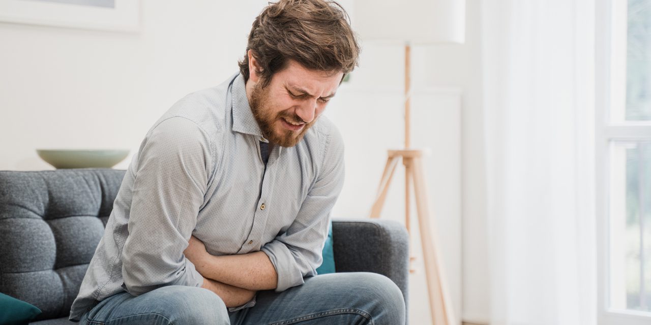 man sitting on sofa holding his belly while showing pain