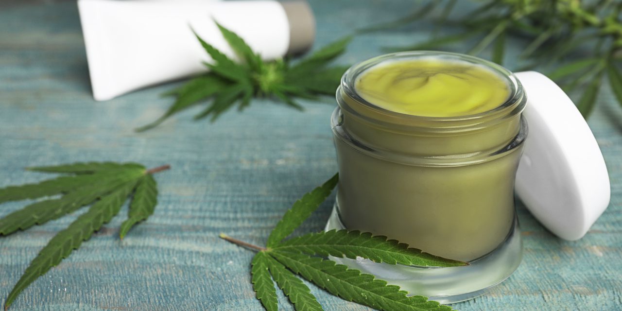 opened cannabis topical in a small jar beside scattered cannabis leaves and topical cream container on the back