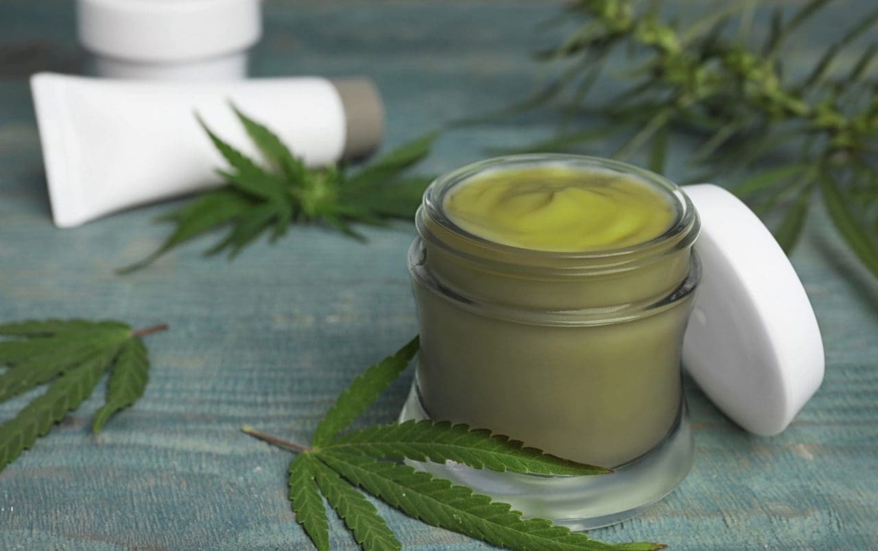 primer plano de cannabis tópico en un pequeño frasco con hojas de cannabis y tubo de crema en el fondo