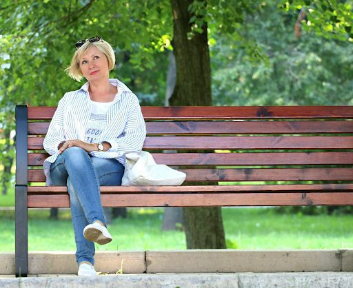 A middle-aged lady sitting on a park bench, near a tree on what looks like a sunny day.