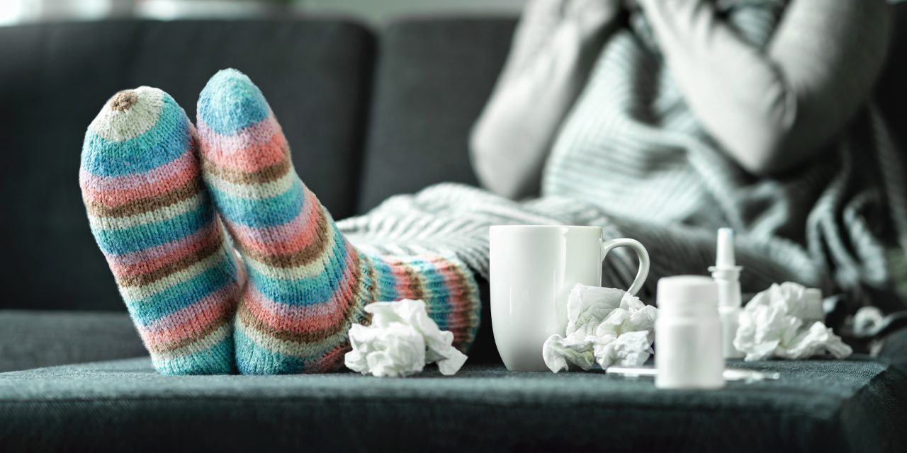 focus of woman's feet wearing colored socks beside a mug, crumpled tissues, medicine container and saline drops